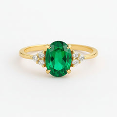 A yellow gold ring featuring a large oval-cut green emerald gemstone in the center, accented by small clear stones on either side of the band, set against a clean white background.