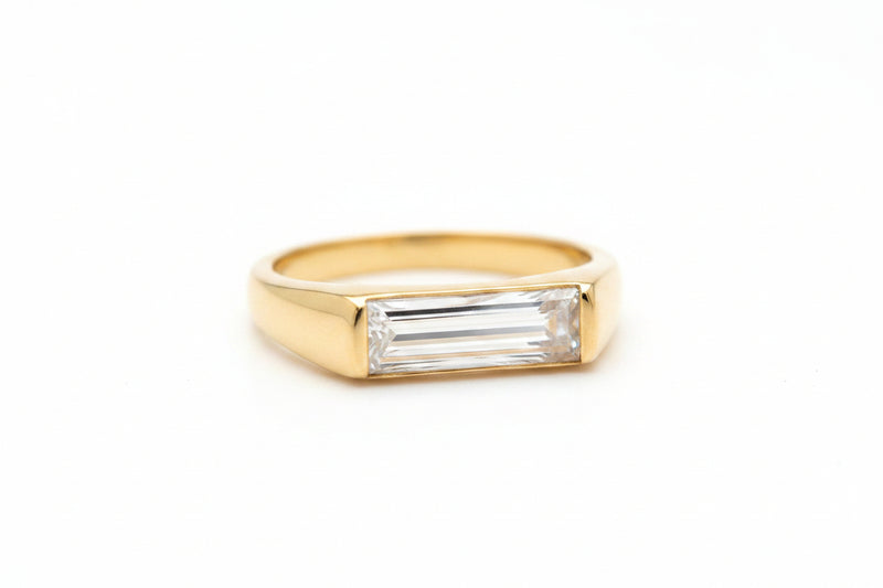 A gold ring with a rectangular baguette-cut clear gemstone, shown worn on skin.
