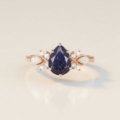 A rose gold ring featuring a central teardrop-shaped dark blue glitter gemstone, flanked by curved rose gold bands with small clear accent stones, set against a light gray reflective background.