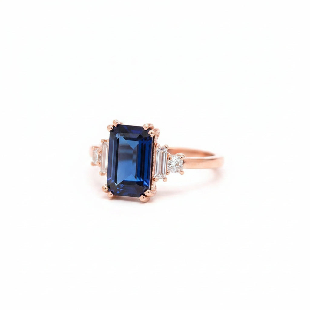 A rose gold ring featuring a rectangular blue sapphire center stone and small clear accent stones on either side, resting on a dark textured stone block with a blurred dark background.