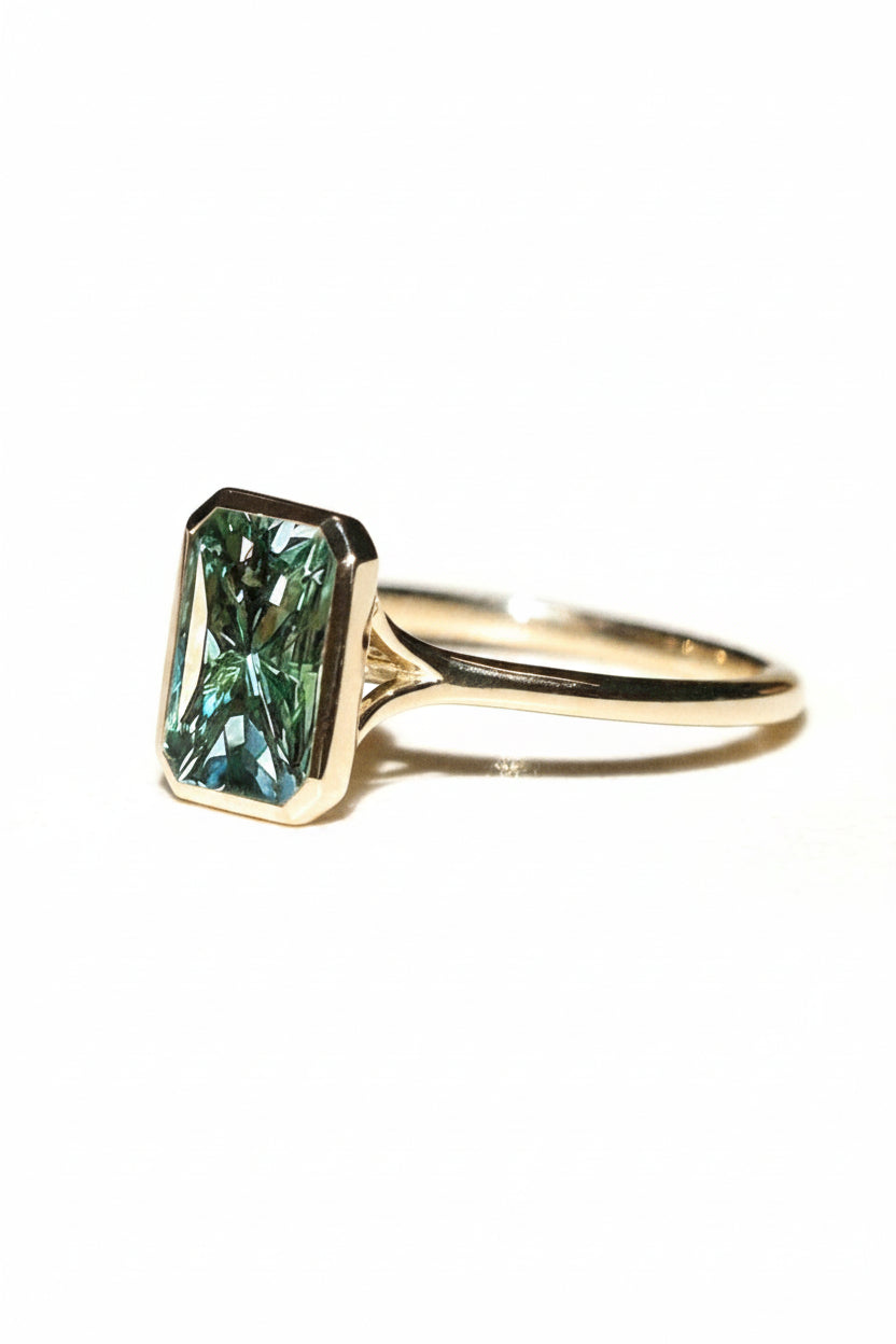 A silver ring featuring a rectangular emerald-cut green gemstone, shown on a plain light beige background.