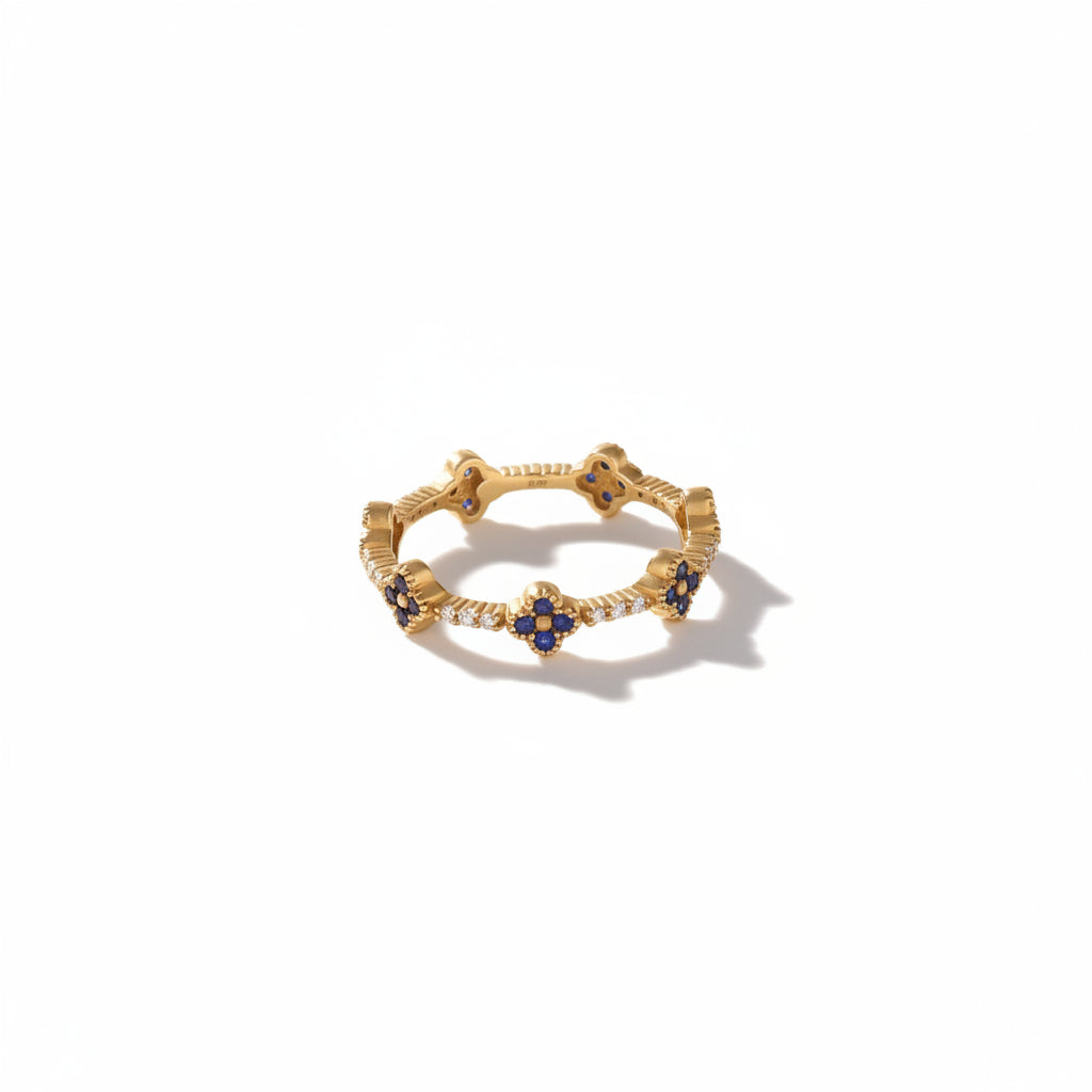 A delicate gold ring with five small blue gemstone clusters spaced evenly around the band, photographed on a warm beige surface with soft shadows and white geometric props in the background.