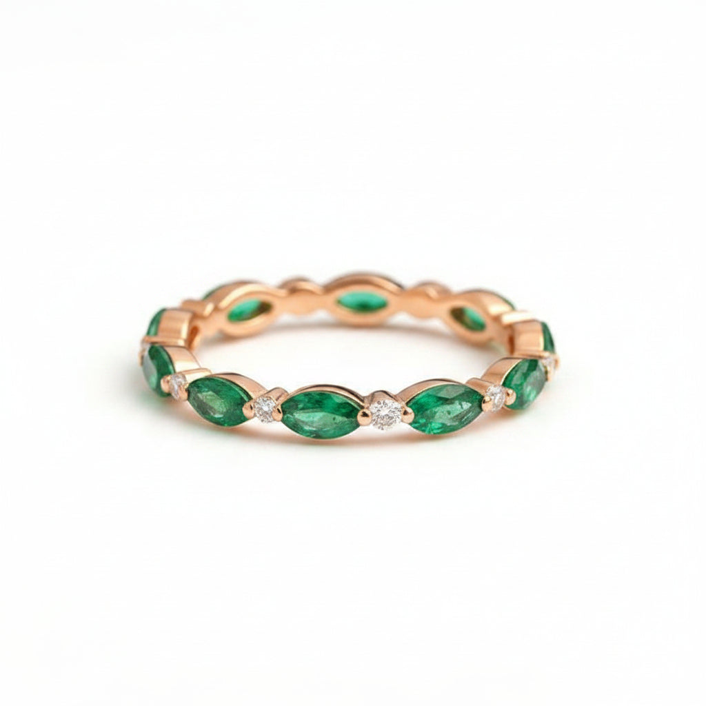 A rose gold eternity band featuring marquise-cut green emeralds and small clear round-cut diamonds, resting on a wooden surface with a lace runner.