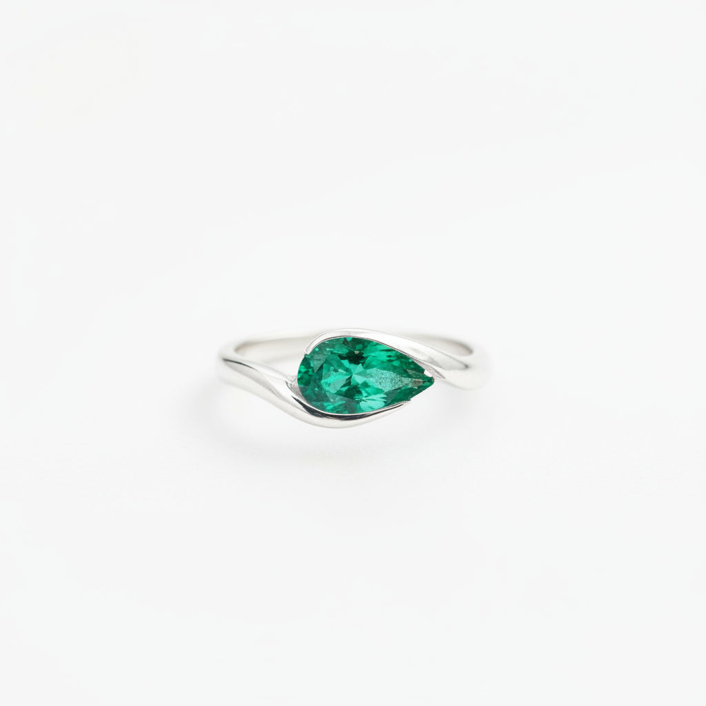A silver ring with a pear-cut green emerald gemstone, resting on textured vintage paper with a lace ribbon and a blurred antique box in the background.