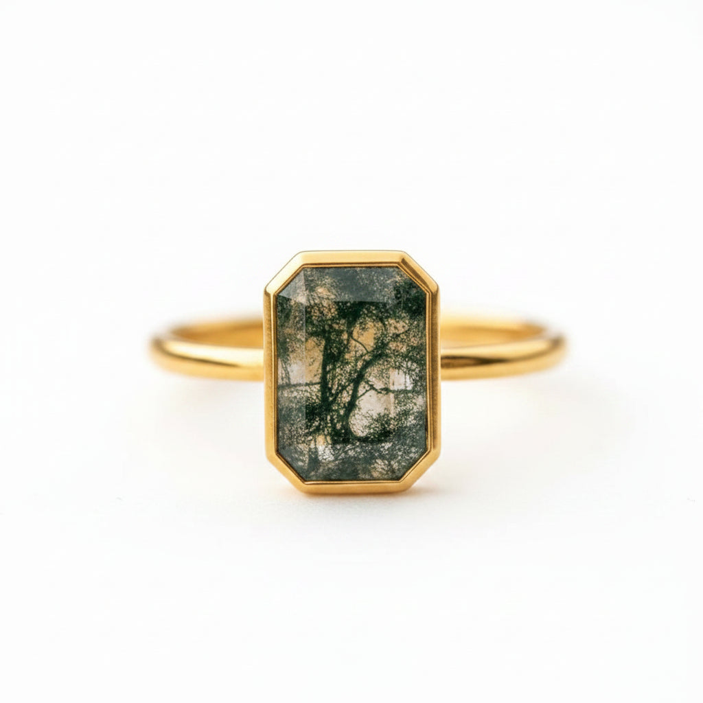 A gold ring with an octagonal bezel holding a dark green and black tree-branch patterned stone, resting on a soft beige fabric surface.