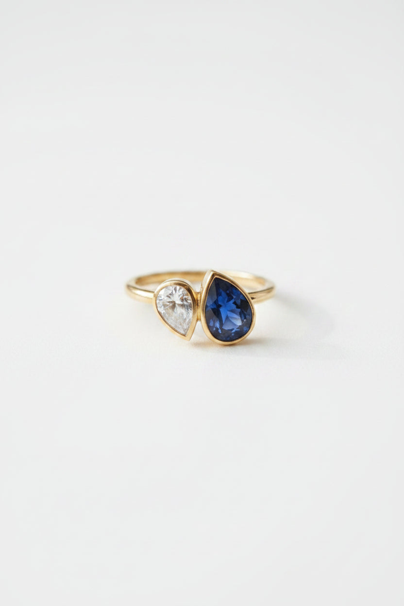 A gold ring with a teardrop-shaped blue sapphire and a smaller clear stone, displayed on an open vintage book with a red wax seal in the background.