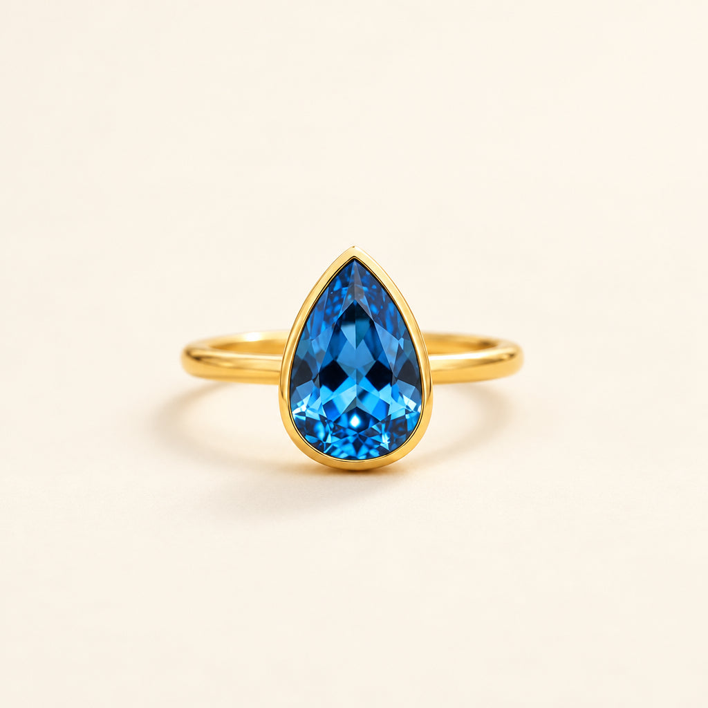 Gold ring with a blue teardrop-shaped gemstone on a soft beige background with dried flowers.