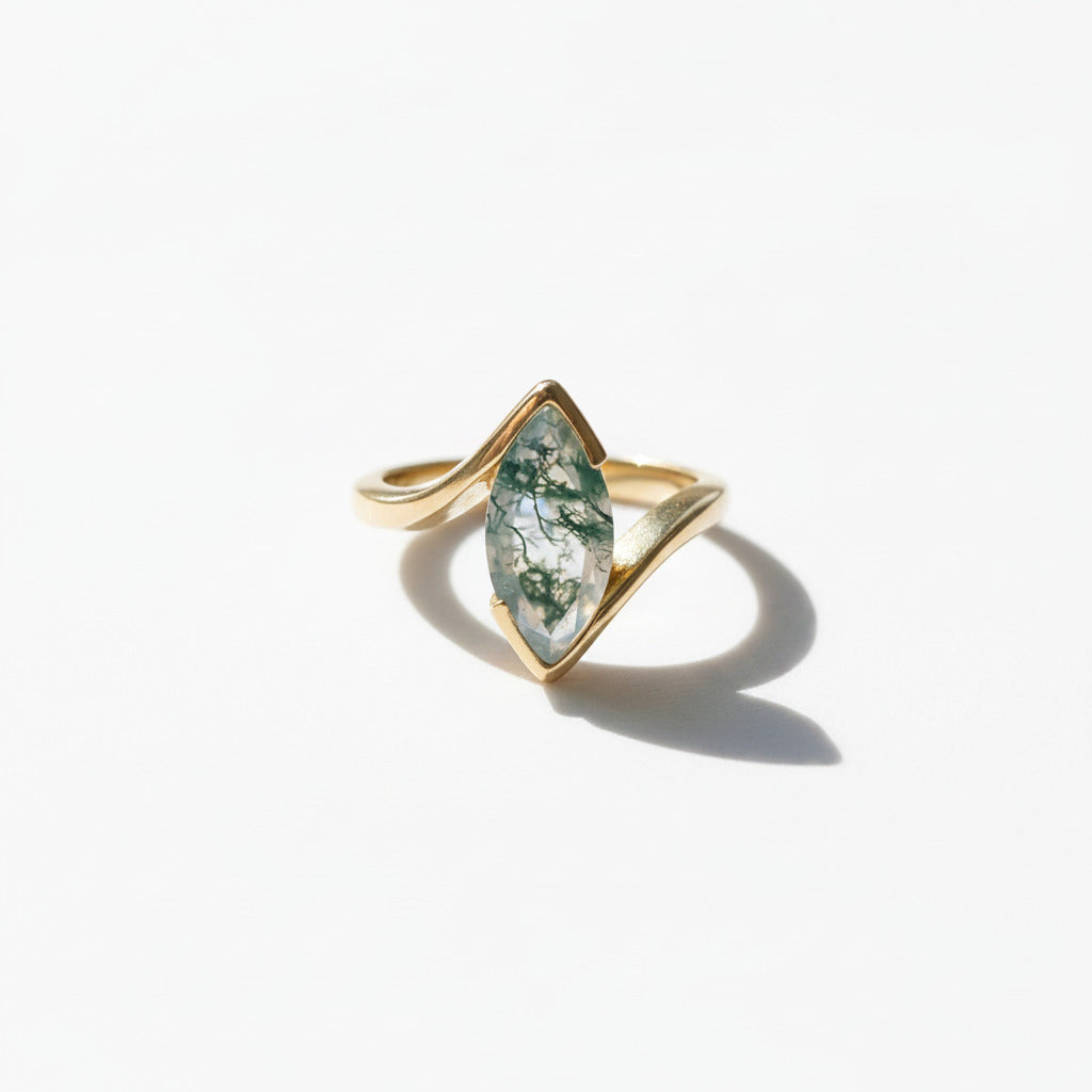 A gold ring with a marquise-shaped green stone containing white inclusions, displayed on a white marble surface with soft shadows.