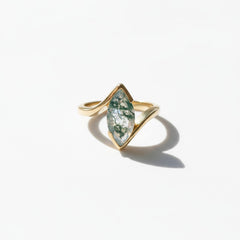A gold ring with a marquise-shaped green stone containing white inclusions, displayed on a white marble surface with soft shadows.