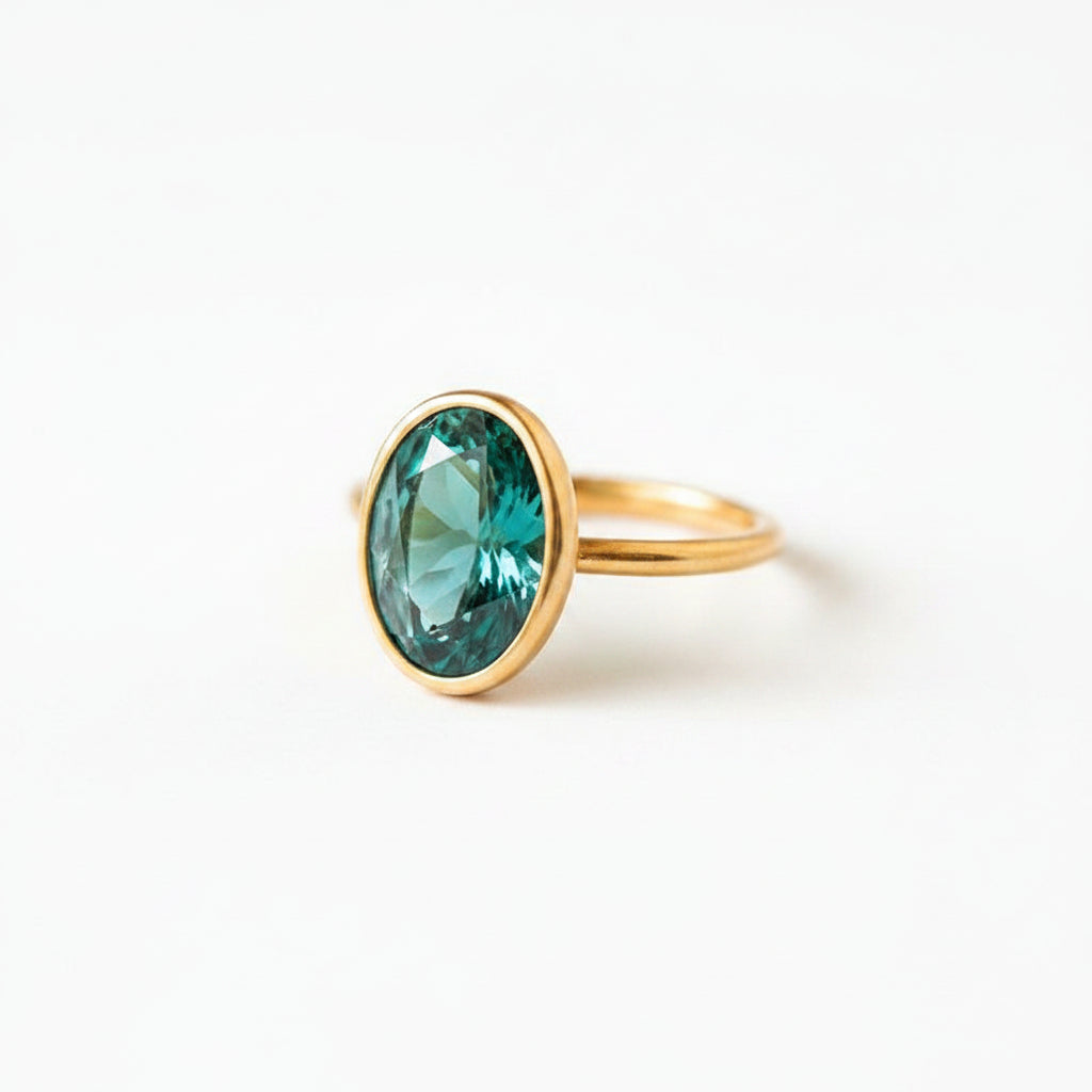 A gold ring with a large oval green gemstone resting on the corner of a brown book with lace fabric nearby.