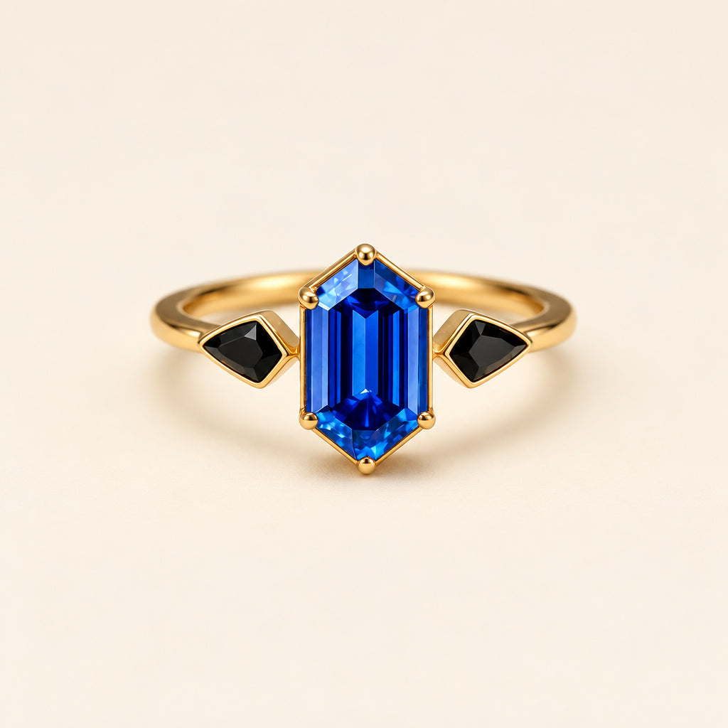 A gold ring featuring a central rectangular blue gemstone and two small black triangular gemstones on either side, displayed on a soft cream fabric background with blurred lace and dried flowers.
