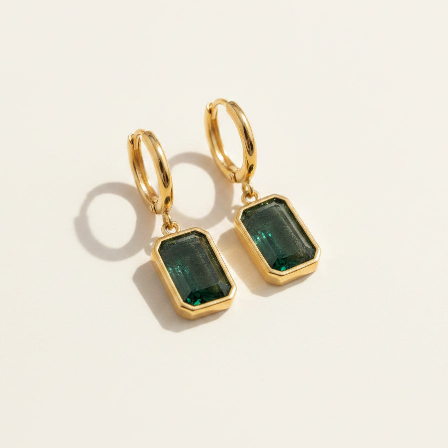 A pair of gold hoop drop earrings with rectangular green gemstone pendants, displayed on a textured gray fabric background.