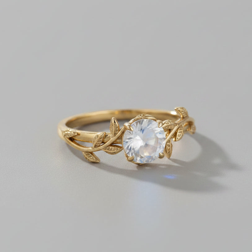 A gold ring with a leafy vine design and a central round iridescent moonstone, resting on a gray surface with soft shadows.