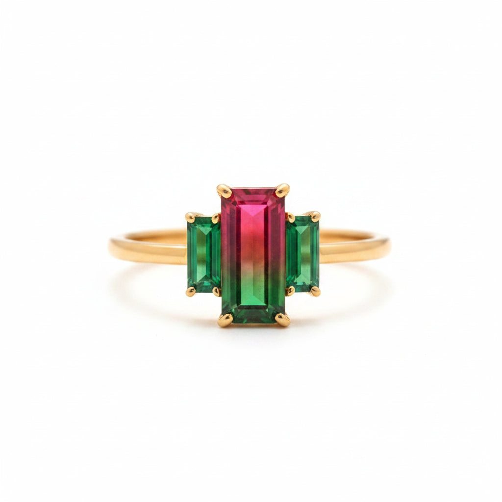 A gold ring with a central rectangular pink-to-green gradient gemstone and two smaller green side stones, displayed on an old wooden book with lace.