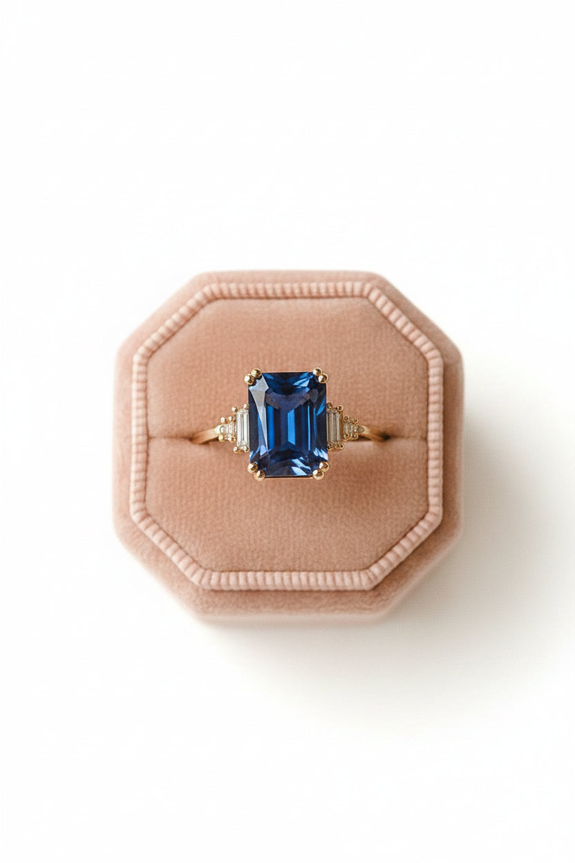 A gold ring with a rectangular blue gemstone displayed on a pink octagonal ring box, resting on a lace doily over a rustic bronze plate.