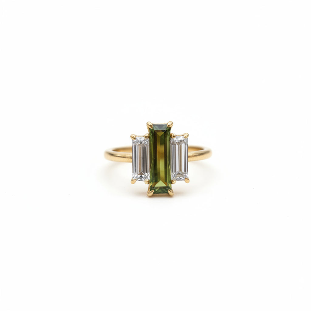 A gold ring with a central green rectangular gemstone and two clear baguette side stones, resting on an old brown book with lace nearby.