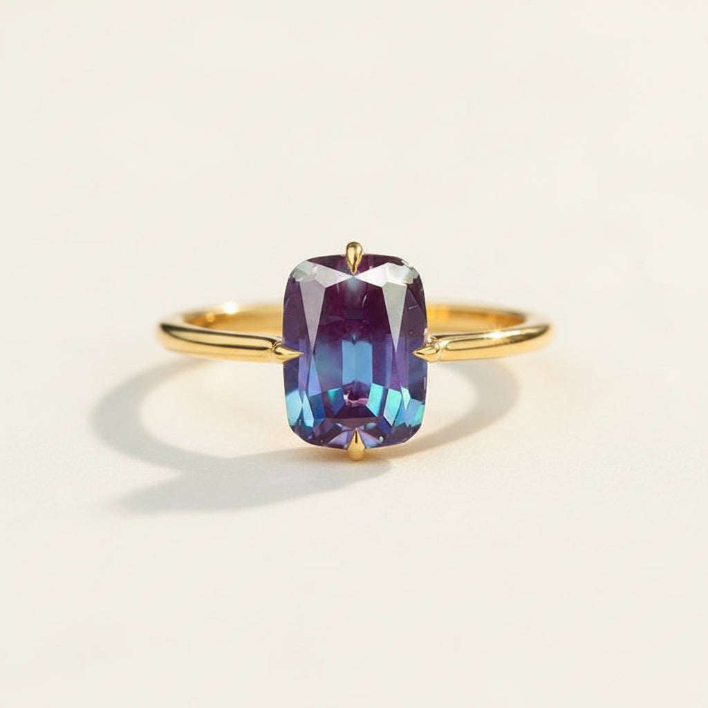 A gold ring with a large rectangular cushion-cut purple gemstone showing blue and violet iridescence, resting on a textured wooden surface with blurred purple lavender in the background.