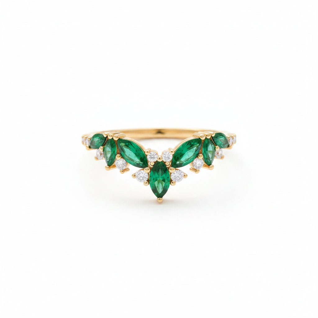 A gold ring with green emerald-cut stones and small clear accent stones, displayed on an old brown book with soft beige fabric nearby.