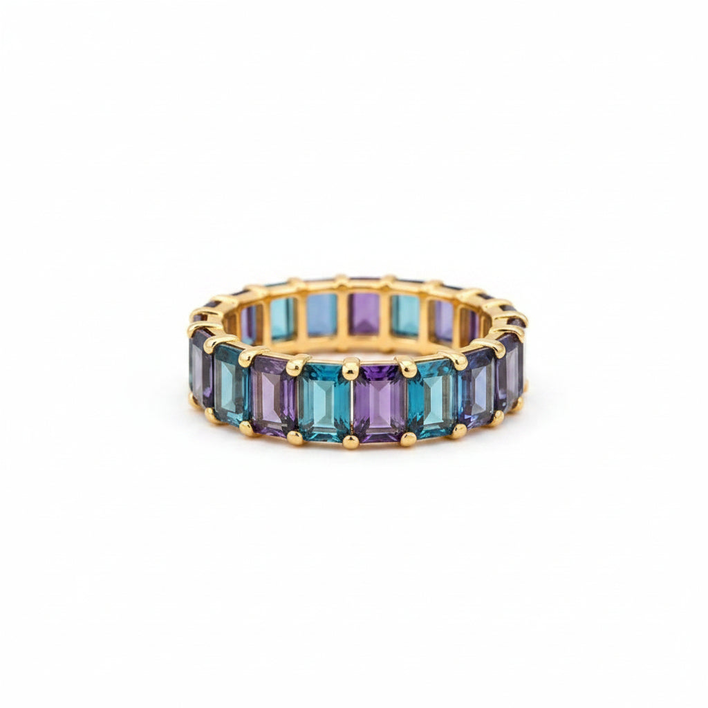 A gold eternity ring with rectangular blue and purple iridescent gemstones, resting on an old brown leather-bound book with lace fabric in the background.