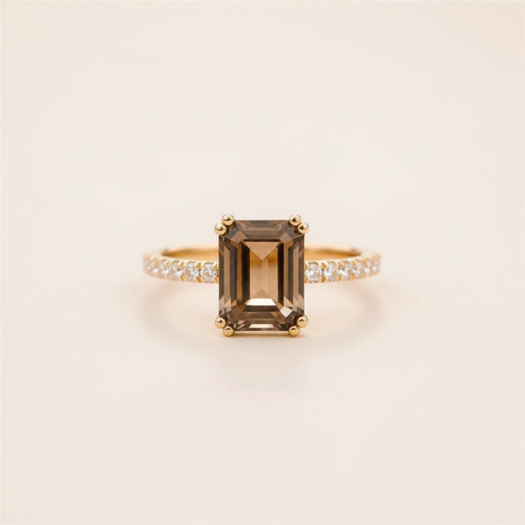 A yellow gold ring featuring a large emerald-cut brown smoky quartz center stone and a pavé-set diamond band, resting on a textured stone surface with a blurred natural background.