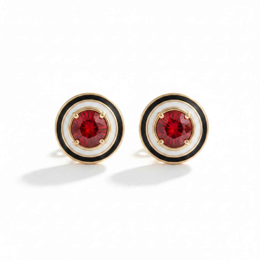 A pair of round stud earrings with gold-tone metal, featuring a white enamel background, a black concentric ring, and a faceted red gemstone in the center, displayed on a textured brown surface.