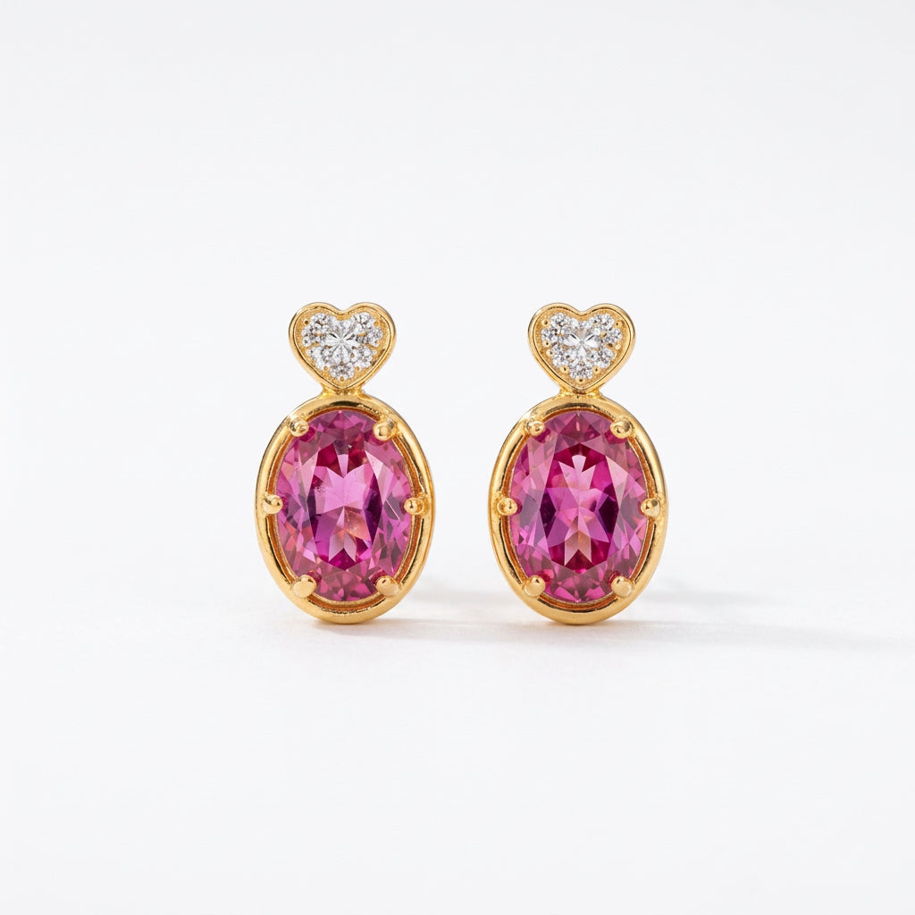 A pair of gold-toned drop earrings with large oval pink gemstones and small heart-shaped clear stones at the top, displayed on a lace fabric against a warm blurred background.