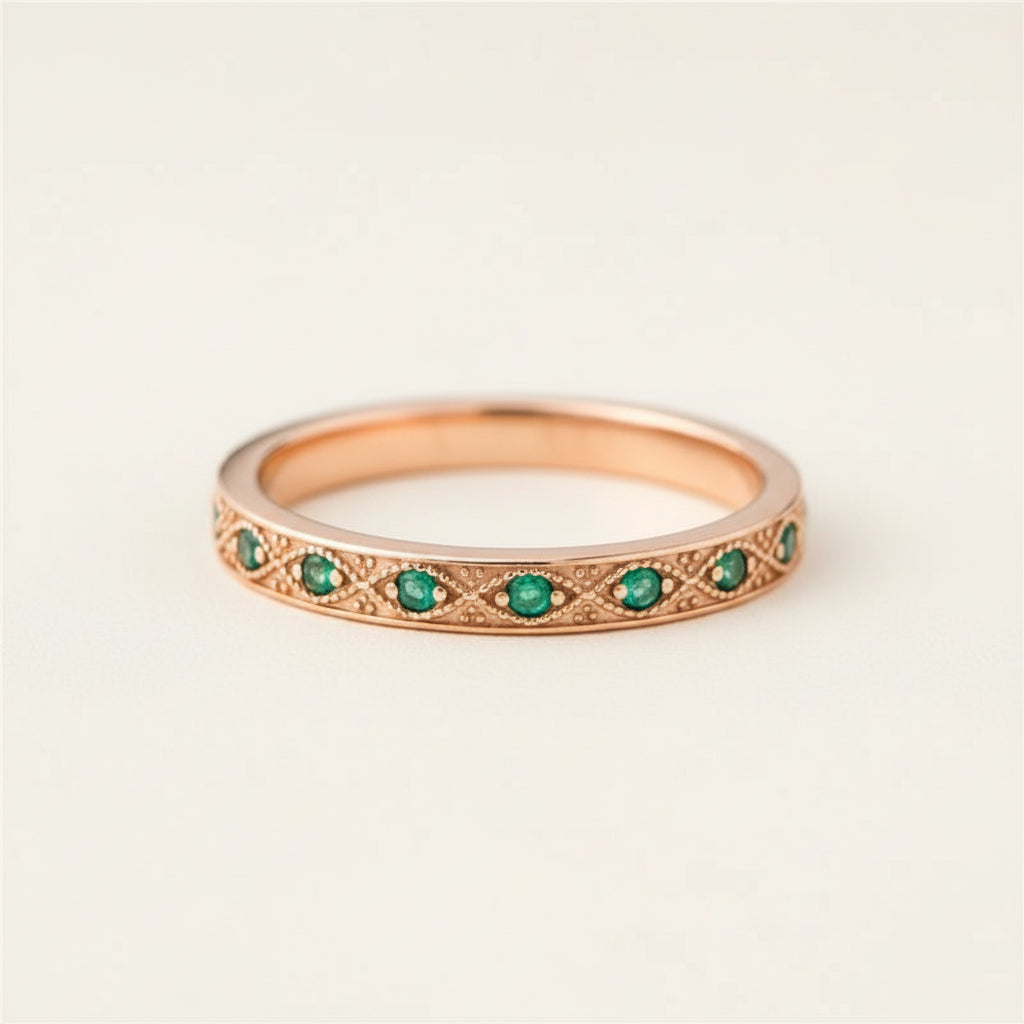 A rose gold eternity ring with round green emerald-cut gemstones, resting on a textured stone surface with blurred purple lavender in the background.