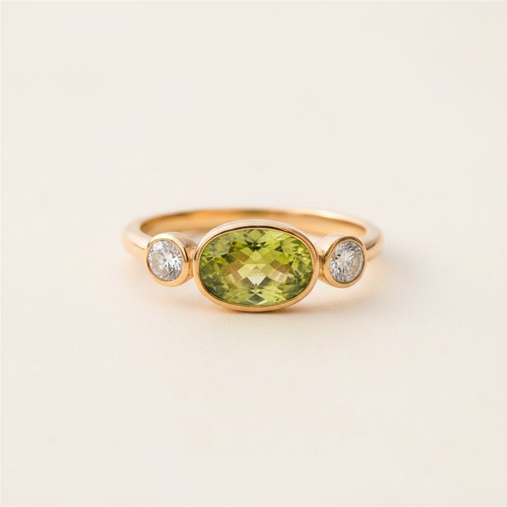 A yellow gold ring featuring a large oval-cut green peridot center stone and two small round-cut clear accent stones on either side, displayed on a textured stone surface with a blurred natural background.
