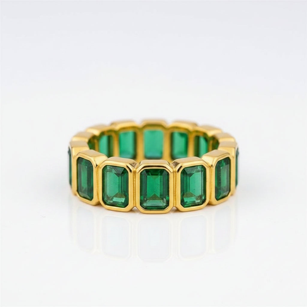 A gold band ring featuring a row of rectangular emerald-cut green gemstones, displayed on a white jewelry box against a dark gray background.