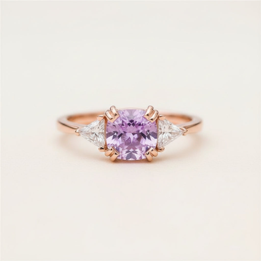 A rose gold ring featuring a central lavender-purple cushion-cut gemstone and two small clear accent stones, resting on a textured stone surface.