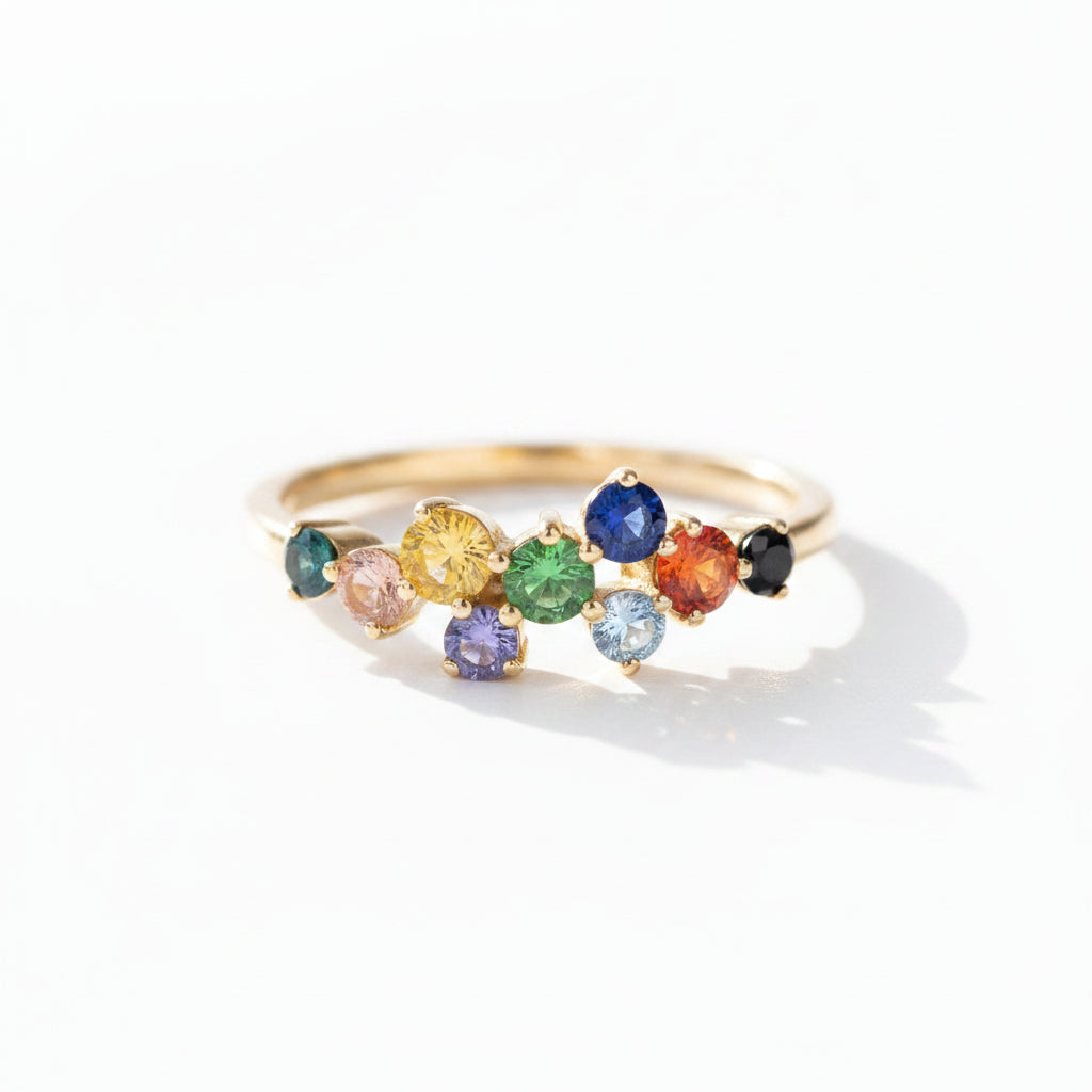 A gold ring featuring a row of colorful gemstones in multiple hues, photographed on a textured gray surface with dramatic sunlight and shadows.