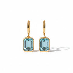 A pair of gold hoop earrings with rectangular light blue gemstones, displayed on a beige stone surface.
