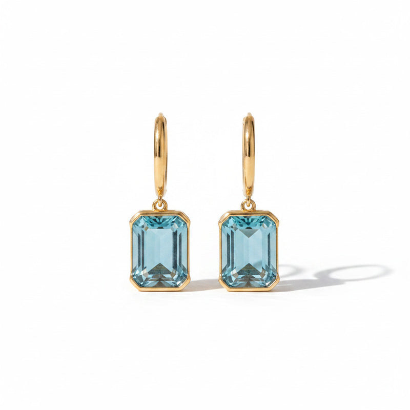 A pair of gold hoop earrings with rectangular light blue gemstones, displayed on a beige stone surface.