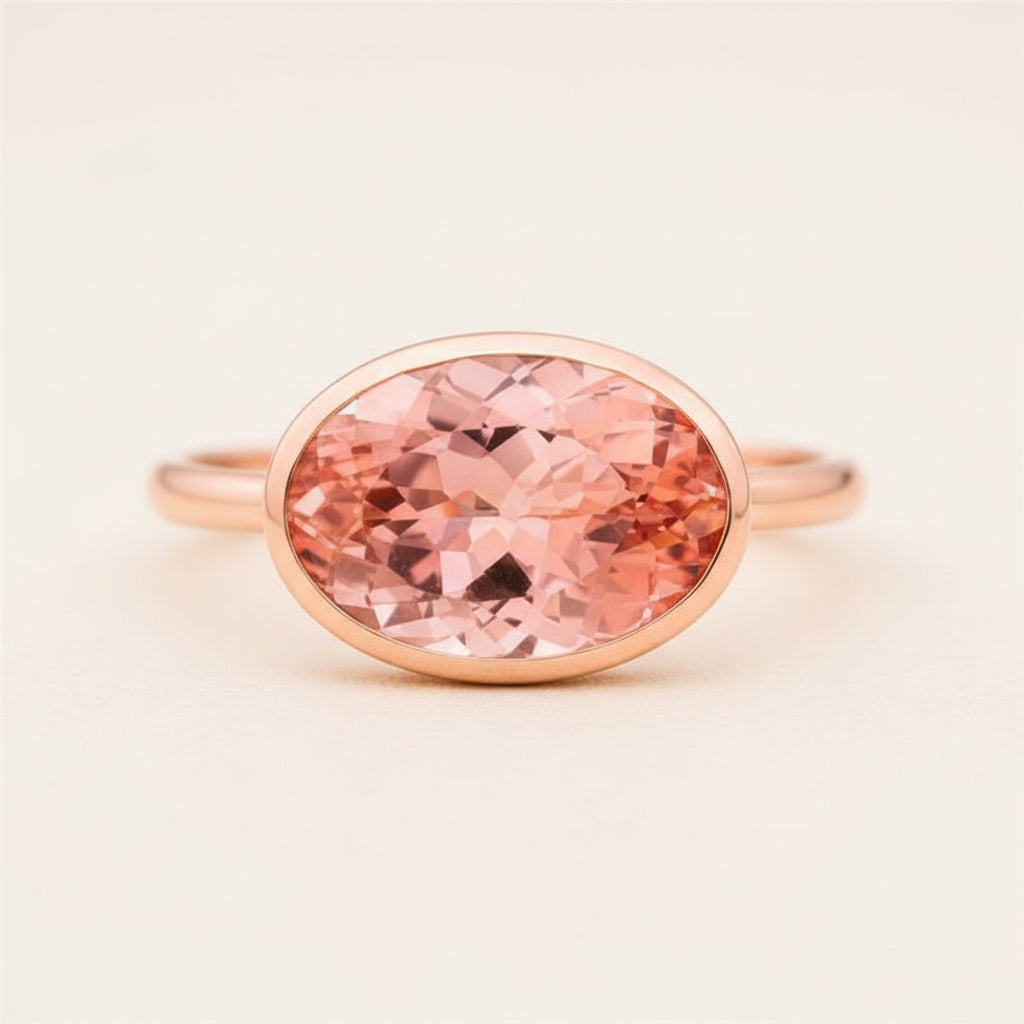 An oval rose gold ring featuring a large, faceted peach-pink gemstone, resting on a textured wooden surface with a soft, blurred natural background.