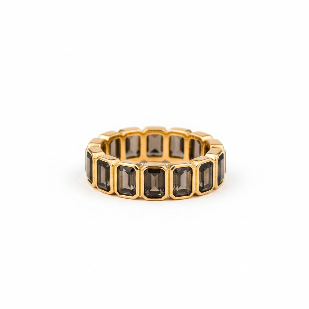 A gold eternity ring featuring rectangular dark smoky gemstones, standing upright on a textured stone surface with a blurred natural background.