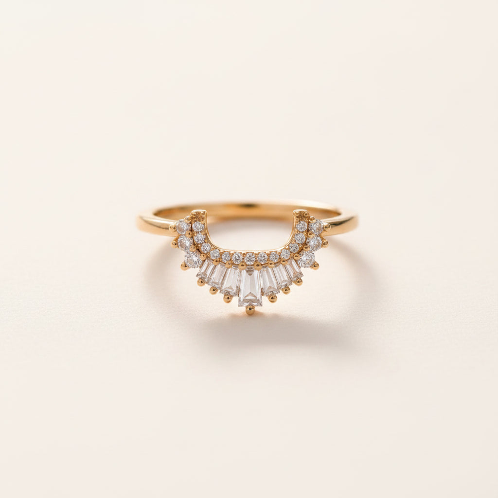 A gold ring with a curved row of clear baguette and round-cut stones, photographed resting on a textured stone surface with a softly blurred natural background.