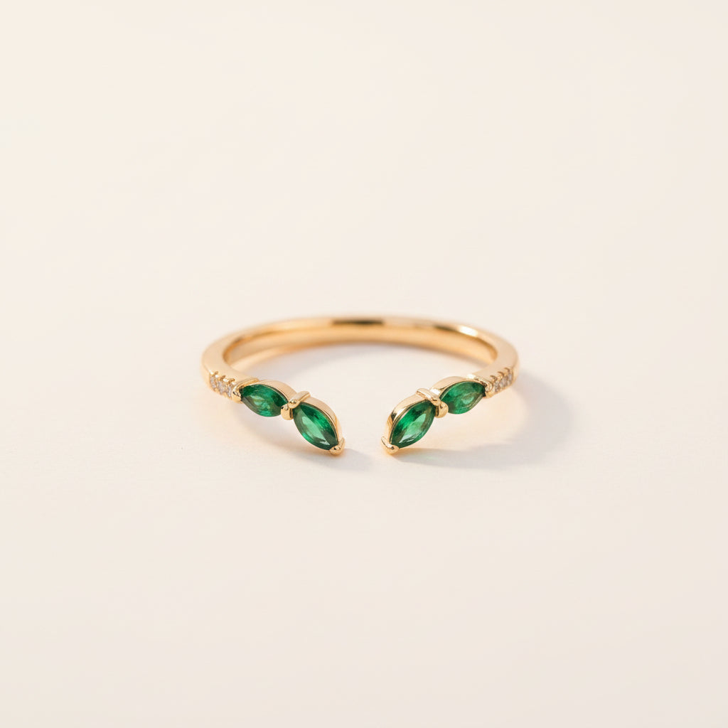 A gold open-bend ring with three marquise-cut green emerald gemstones and small clear accent stones, resting on a textured stone surface with blurred green foliage in the background.