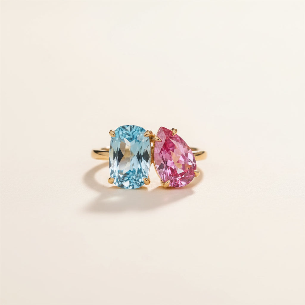 Two yellow gold rings, each featuring a large gemstone, resting on a textured stone surface. The ring on the left has a cushion-cut light blue gemstone, and the ring on the right has a pear-cut pink gemstone.