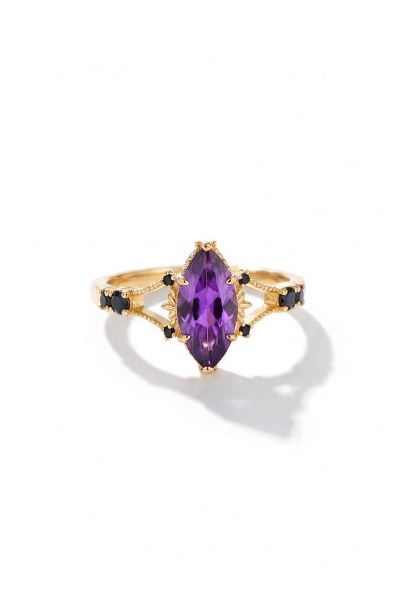 A gold ring with a large marquise-cut purple amethyst center stone and small black accent stones on the band, displayed on a gray surface with a dark background.