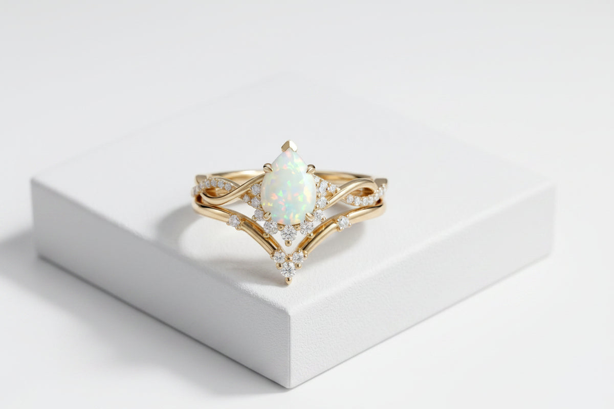 A gold ring with a pear-shaped opal center stone and small clear accent stones, displayed on a white rectangular pedestal against a gray textured background.
