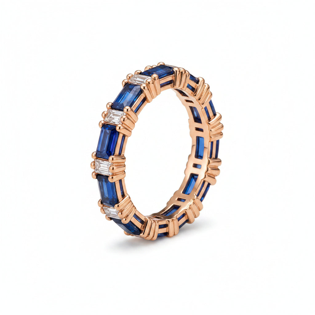 A rose gold eternity ring featuring baguette-cut blue sapphires and small clear accent stones, displayed on an antique brown book next to a beige lace cloth.