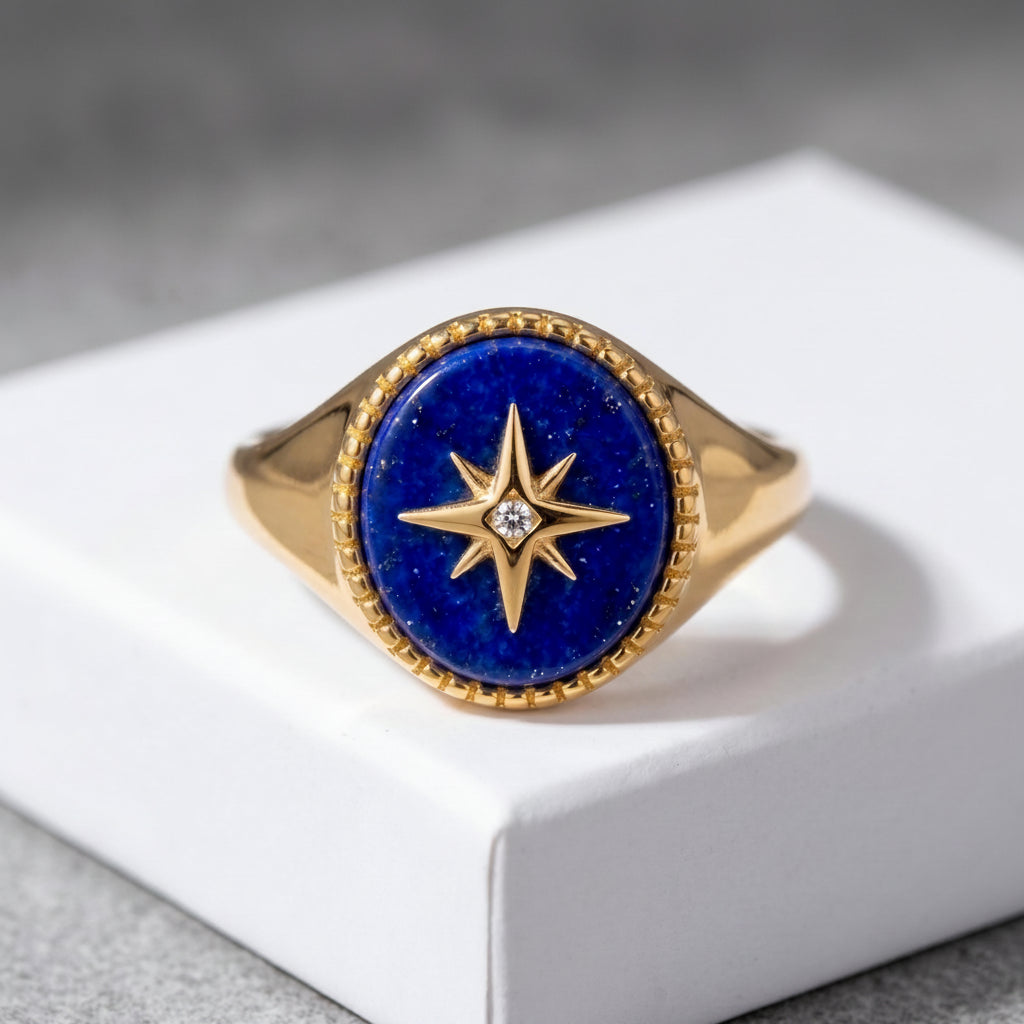 A gold signet ring with a deep blue lapis lazuli stone face and a gold compass star motif, resting on a white display block.