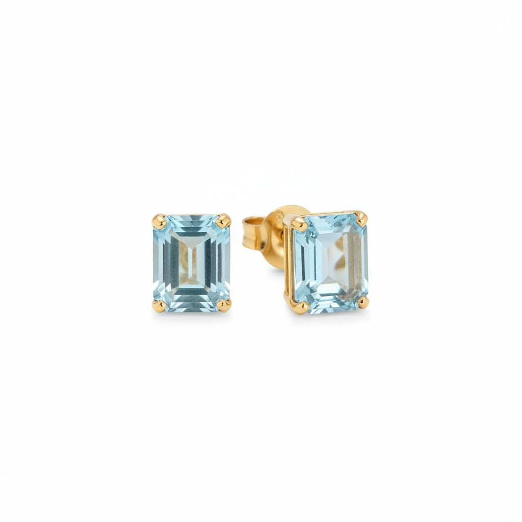 A pair of gold stud earrings with rectangular light blue gemstones, resting on a polished wooden surface.
