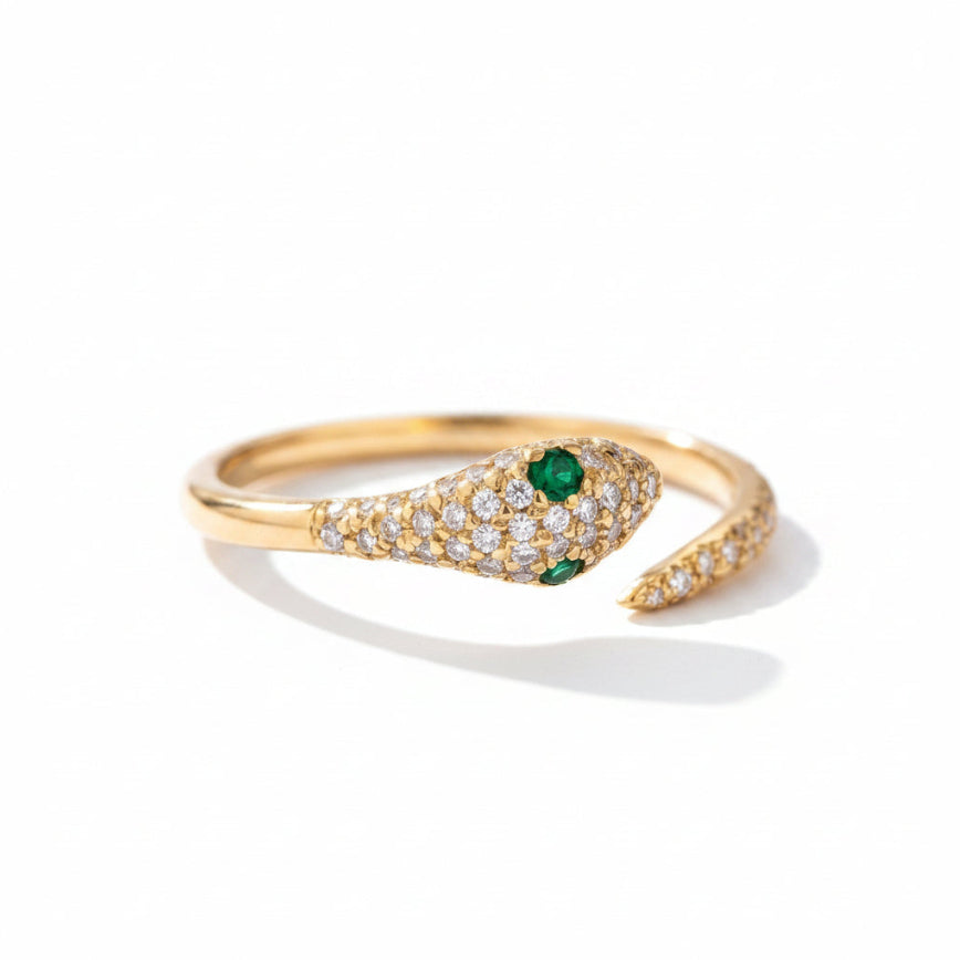 A gold snake ring with pavé-set clear stones and two green emerald-cut gemstones, photographed on a dark textured surface.