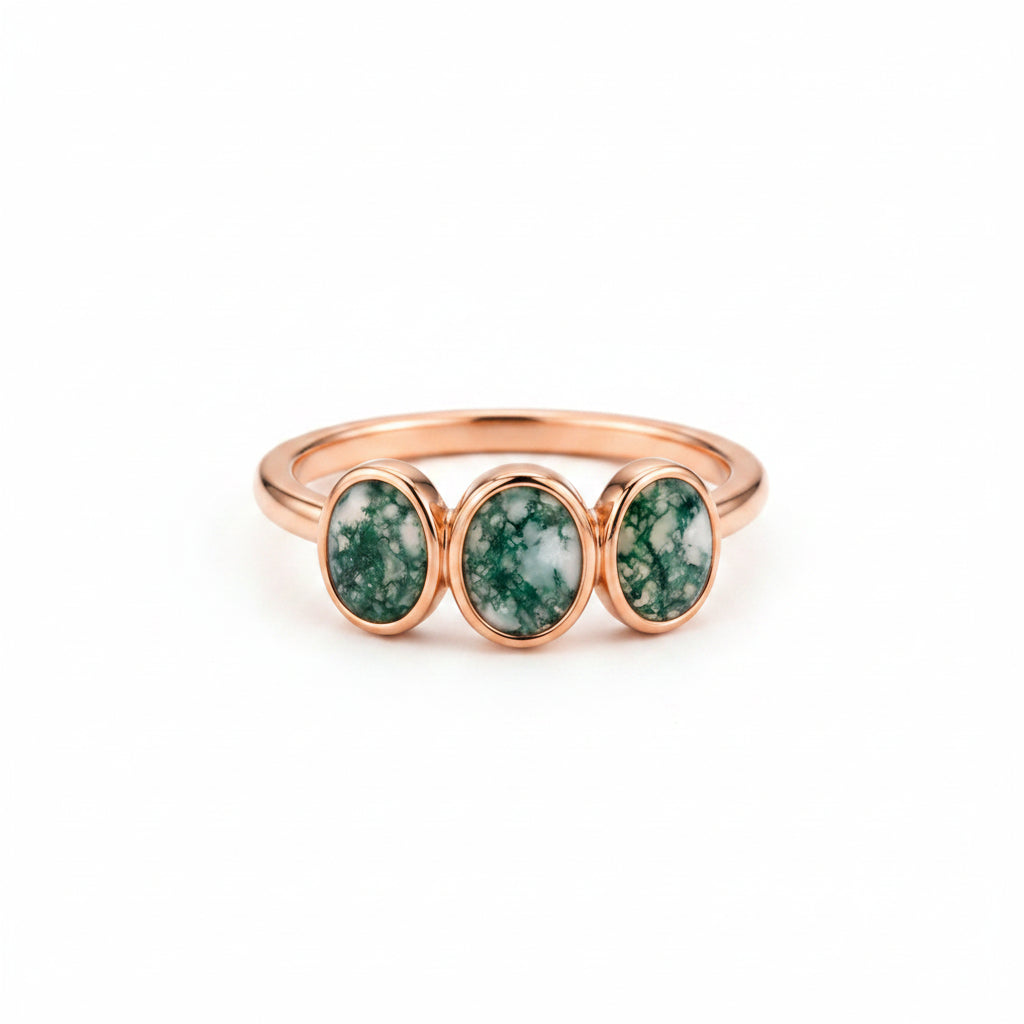 A rose gold ring with three oval green and white marbled gemstones, resting on an antique brown book with lace draped over it.