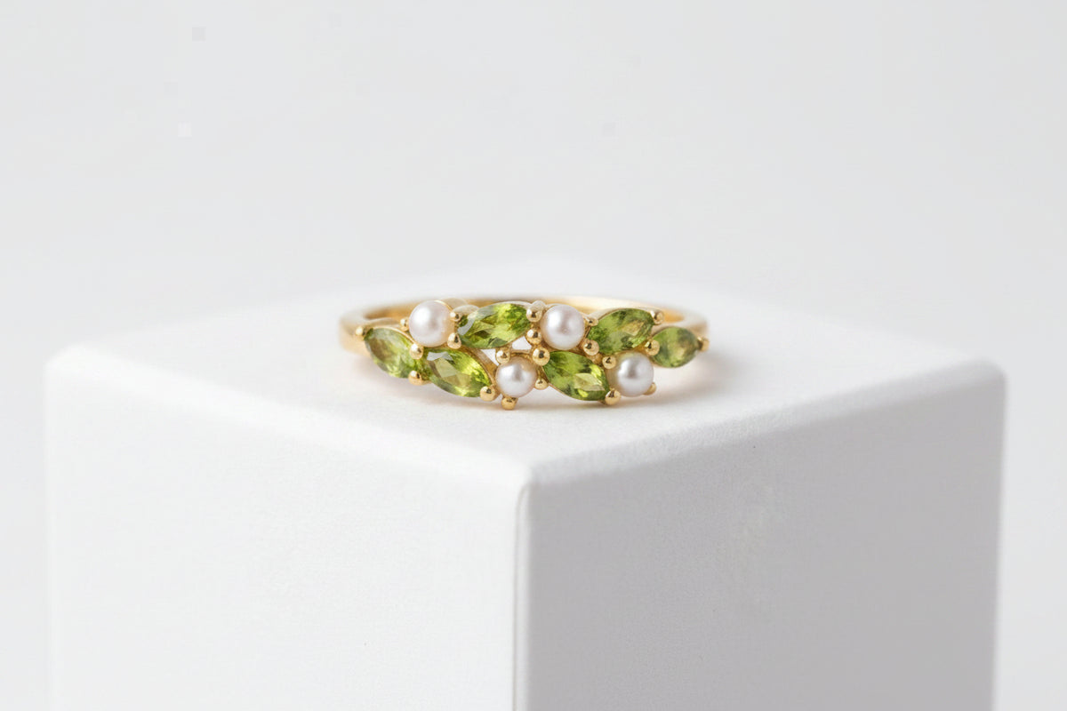 A gold ring featuring marquise-cut green peridot stones and three white pearls, displayed on a white geometric block against a soft gray background.