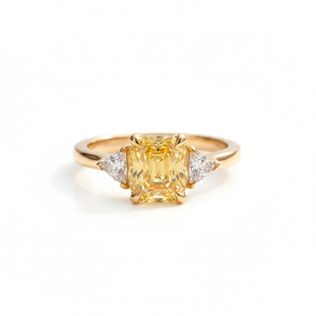 A yellow gold ring with a large cushion-cut yellow center stone and two small clear side stones, displayed on textured cream fabric with soft green leaves blurred in the background.