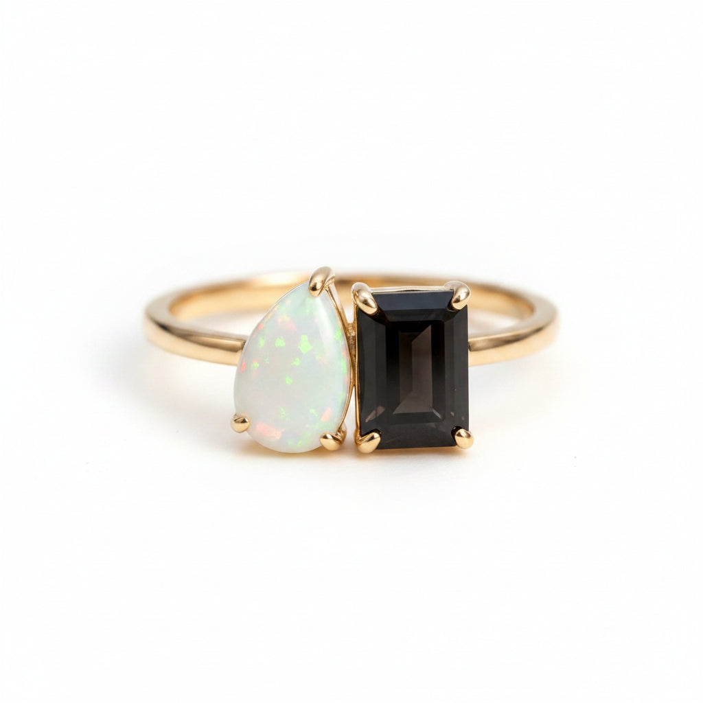 Gold ring with a teardrop-shaped opal and rectangular smoky stone on a textured surface with green leaves in the background.