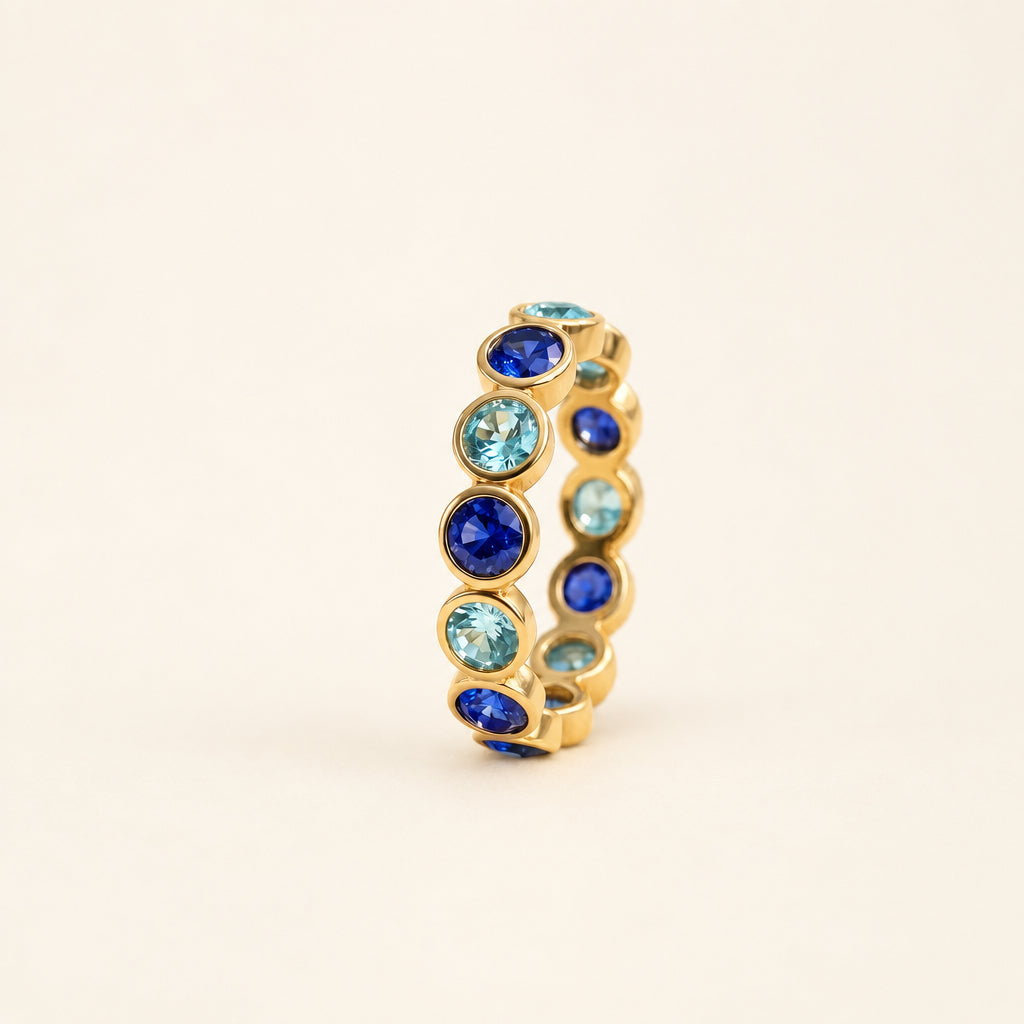 A gold band ring featuring alternating round blue and light aqua gemstones, resting on a textured beige lace fabric with soft green leaves blurred in the background.