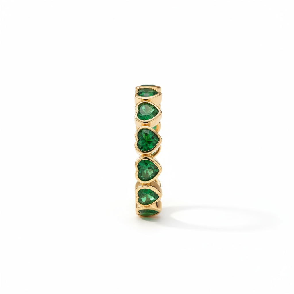 A gold ring featuring four heart-shaped green emerald gemstones standing upright on a bed of sand, styled with a piece of beige linen and eucalyptus leaves.
